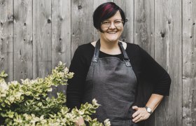Wirtin Petra Haselsteiner, © Niederösterreich Werbung/David Schreiber Eine Frau mit Brille und Schürze steht lächelnd vor einer Holzwand neben einem Busch.