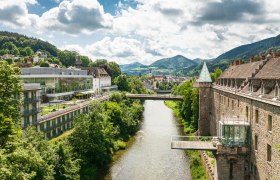 Das Schloss an der Eisenstrasse, © Dominik Stixenberger Panorama einer Flusslandschaft mit Schloss und moderner Architektur, umgeben von grünen Hügeln.