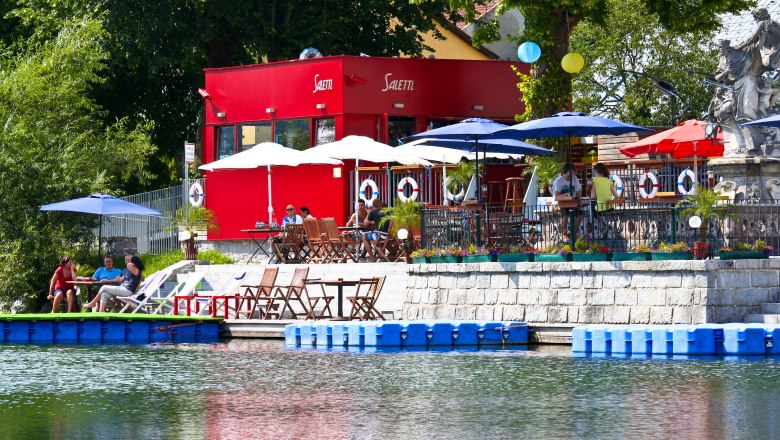 Salettl, © zVg Ein rotes Gebäude mit Terrasse am Wasser, umgeben von Sonnenschirmen und Sitzgelegenheiten.