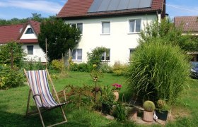 Hausansicht mit Garten, © Familie Maier Ein Haus mit Solarpanelen auf dem Dach, umgeben von einem Garten mit Pflanzen und einem Liegestuhl.