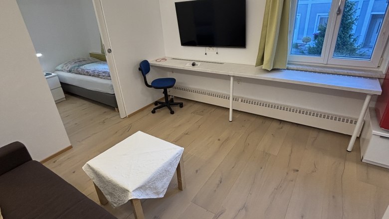 TDR-Haus Morgensonne Wohnzimmer, © Fam Pauker Modernes Wohnzimmer mit braunem Sofa, kleinem Tisch, Fernseher an der Wand, Schreibtisch und Stuhl. Fenster mit Blick auf ein blaues Gebäude.