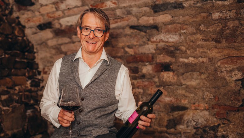 Wirt und Weinexperte: Martin Teufl, © Niederösterreich Werbung/Daniela Führer Ein Mann mit Brille hält ein Weinglas und eine Weinflasche vor einer Ziegelwand.