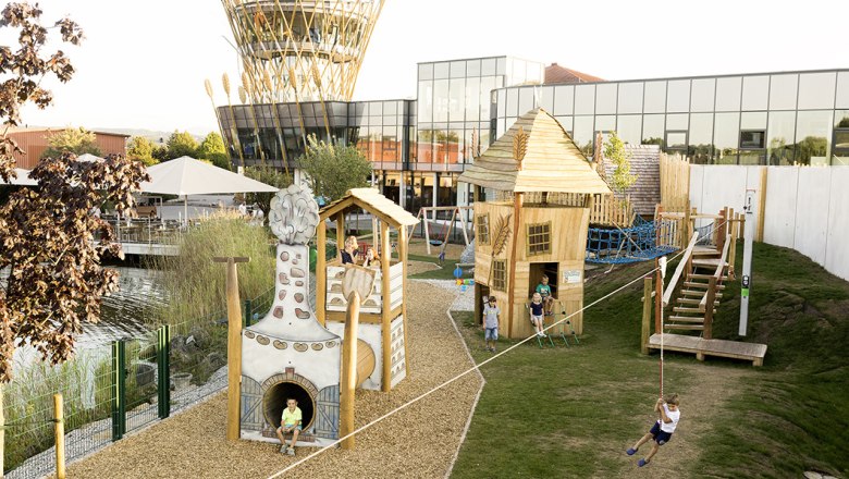 Haubiversum playground, © Haubis GmbH A modern playground with wooden houses and a rope slide in front of a building with a glass façade and a tower with wheat ornaments.