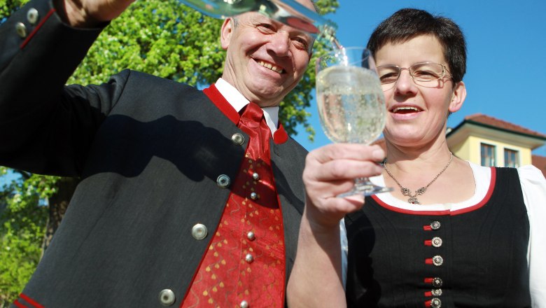 Mostbaron Wieser, © Mostviertel Tourismus, weinfranz.at Zwei Personen in traditioneller Kleidung stoßen mit einem Glas an, während eine Flasche eingeschenkt wird.