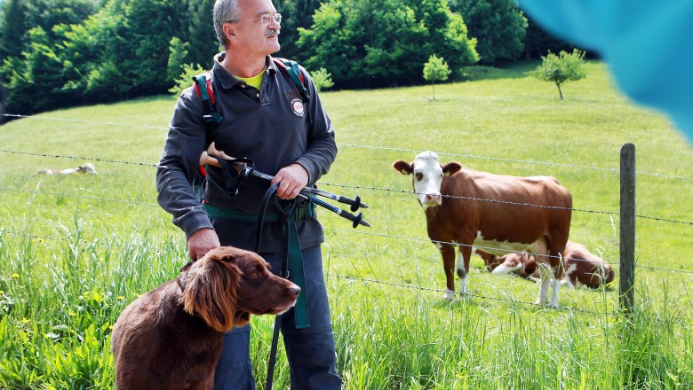 Hiking around the Muckenkogel, © weinfranz.at A man with a dog and walking sticks stands in front of a pasture with cows.