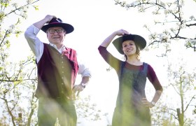 Distelberger, © Martina Schuller Zwei Personen in traditioneller Kleidung mit Hüten stehen in einem blühenden Obstgarten.