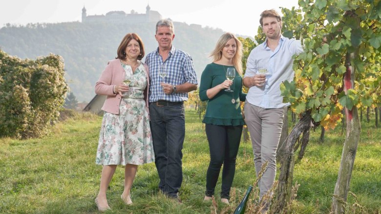 Familie Heinz Fink, © Philipp Monihart Vier Personen stehen in einem Weinberg mit Weingläsern in der Hand, im Hintergrund ist eine Burg auf einem Hügel zu sehen.