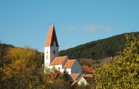Pfarrkirche Nußdorf, © Blesl Pfarrkirche Nußdorf mit rotem Dach und Uhrturm, umgeben von Bäumen und Hügeln.