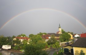 Regenbogen über Gerersdorf, © Schild Renate Ein Regenbogen spannt sich über ein Dorf mit Kirche und Häusern in Gerersdorf.