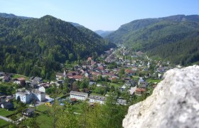 Blick von der Burgruine, © Gemeinde Hohenberg Panoramablick auf ein Dorf in einem grünen Tal, umgeben von bewaldeten Hügeln, mit einem Felsen im Vordergrund.