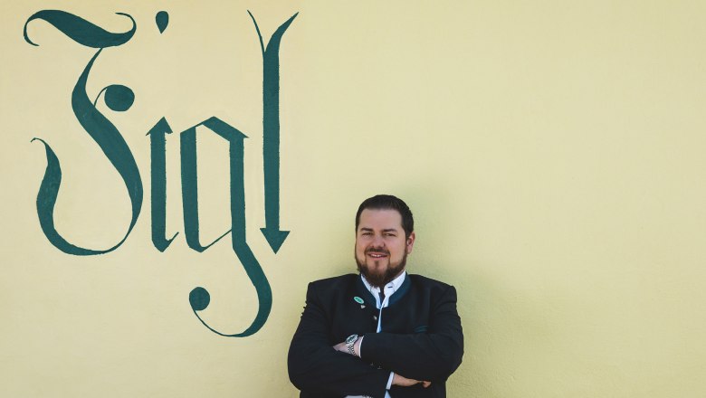 Matthias Strunz, der Wirt vom Figl, © Niederösterreich Werbung/David Schreiber Ein Mann in traditioneller Kleidung steht vor einer gelben Wand mit dem Schriftzug 'Figl'.