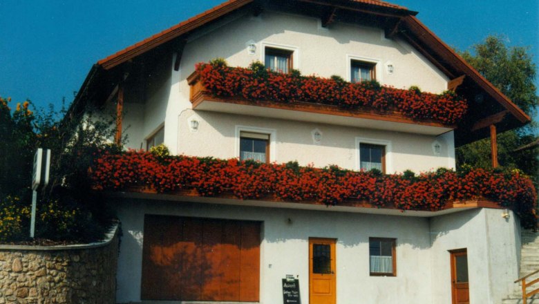 Gästehaus Huber, © Gästehaus Huber Ein traditionelles Gästehaus mit roten Blumen auf den Balkonen und einem roten Dach.