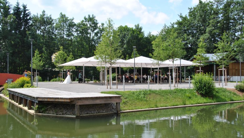 Gemeinde Zeillern, © zVg Gemeinde Zeillern Ein Hochzeitspaar steht vor einem Pavillon mit Gästen, umgeben von Bäumen und einem Teich.