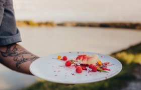 Wolke 7: Weißes Schokomousse mit Himbeergelee & Himbeerstaub, © Niederösterreich Werbung/Daniela Führer Ein Teller mit weißem Schokomousse, Himbeeren und Blüten, gehalten von einer tätowierten Hand vor einem See.