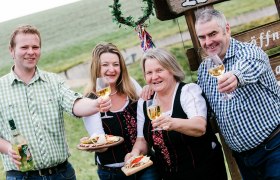 Mostheuriger z'Gridling, © schwarz-koenig.at Vier Personen stoßen mit Weingläsern an, im Freien vor einem Schild.