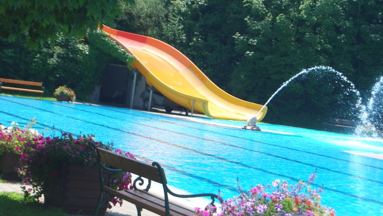 St. Valentin outdoor pool, © zVg Gemeinde St. Valentin An outdoor pool with a yellow slide, water fountain and flowers in the foreground.