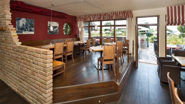 Gastzimmer, © DR Ein gemütliches Restaurant mit Holzmöbeln, roten Wänden und großen Fenstern, die auf eine Terrasse führen.