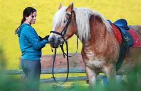 Schnupperreiten am Bodenhof, © weinfranz.at Eine Frau steht neben einem gesattelten Pferd und hält die Zügel. Im Hintergrund ist eine gelbe Wand zu sehen.