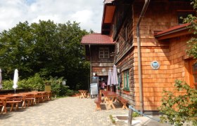 Hauptansicht der Kaiserkogelhütte, © ÖTK Eschenau Holzhütte mit Terrasse und Sitzbänken im Freien.