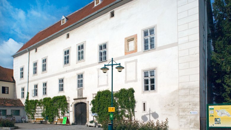 Schloss Traismauer, © © Stadtgemeinde Traismauer-Marius-Höfinger Schloss Traismauer mit weißer Fassade und roten Dachziegeln, umgeben von Pflanzen und einem gepflasterten Vorplatz.