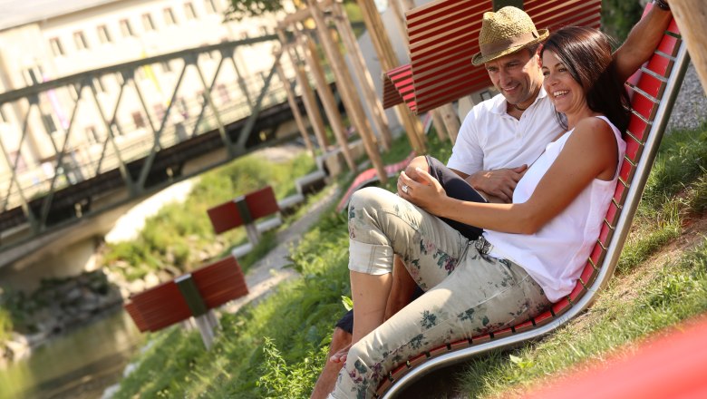 Erlaufhafen Begegnungszone, © schwarz-koenig.at Ein Paar sitzt entspannt auf einer roten Liege im Freien, nahe einer Brücke.