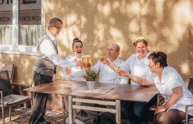 Starkes Wirtshaus-Team, © Niederösterreich Werbung/Daniela Führer Ein Team von fünf Personen sitzt draußen an einem Tisch und stößt mit Getränken an. Ein schwarzer Hund sitzt davor.