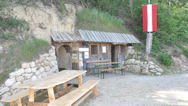 Cave, © ZVG Gemeinde A rustic wooden hut with a table and benches in front of it, built into a hill. A red and white flag hangs from a tree.