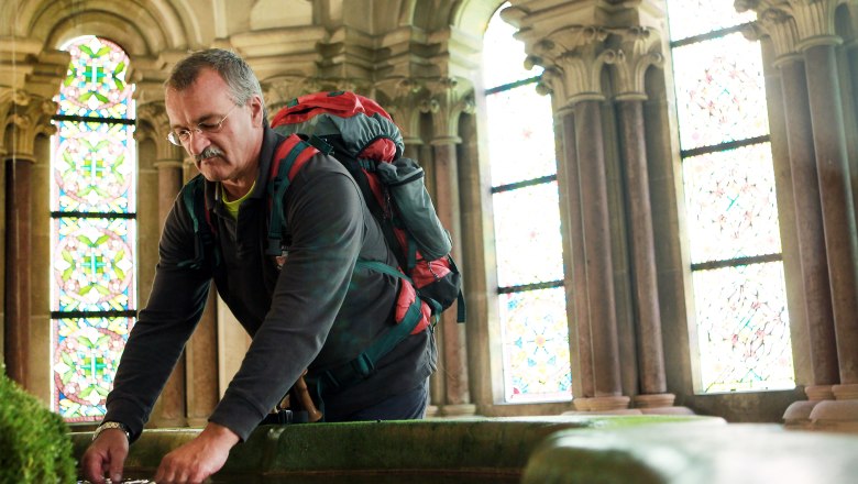 Stift lilienfeld, © Mostviertel Tourismus/Weinfranz.at A man with a rucksack bends over a pool of water in a historic building with stained glass windows.