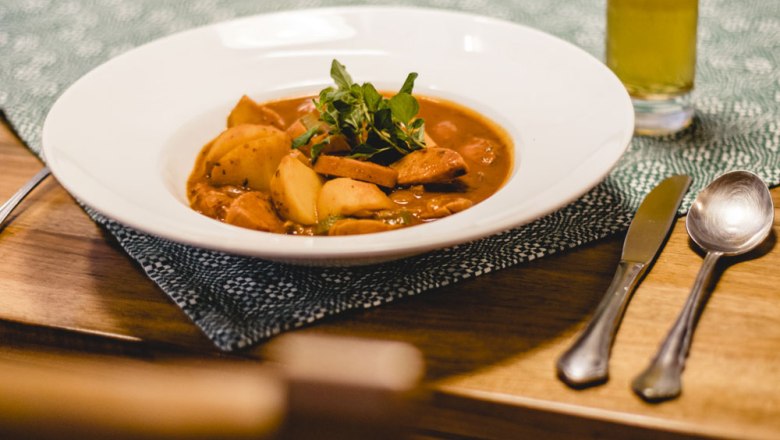 Kartoffelgulasch, © Niederösterreich Werbung/David Schreiber Ein Teller mit Kartoffelgulasch auf einem Tisch, daneben Besteck und ein Glas Getränk.