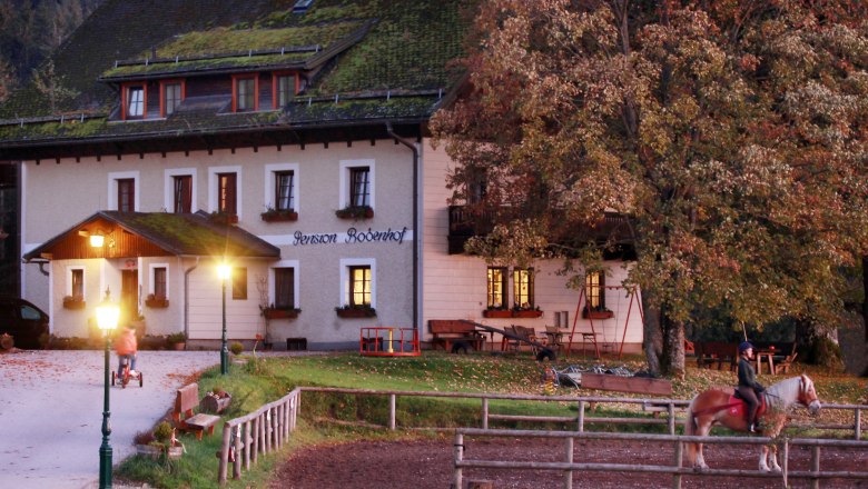 Vacation on the farm at Bodenhof, © weinfranz.at A farm with illuminated windows, a rider on a horse and a child on a tricycle in the foreground.