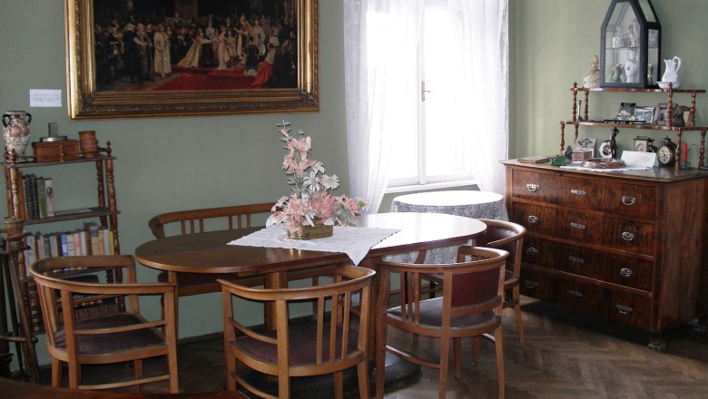 Heim, © Stadtgemeinde Mank Ein traditionell eingerichtetes Wohnzimmer mit Holzmöbeln, einem runden Tisch, Stühlen, einer Kommode und einem großen Gemälde an der Wand.