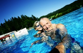 Freibad Mitterbach, © weinfranz.at Ein älteres Paar schwimmt lächelnd in einem Freibad mit Wald im Hintergrund.