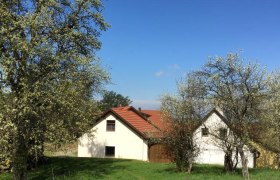 Hochbrand, © zVg Ein weißes Haus mit rotem Dach steht auf einer grünen Wiese, umgeben von blühenden Bäumen unter einem klaren blauen Himmel.