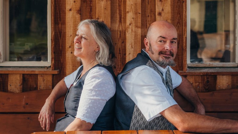 Innkeepers Renate and Petter Kummer, © Niederösterreich Werbung/Daniela Führer A man and a woman sit back to back on a wooden bench in front of a wooden building.