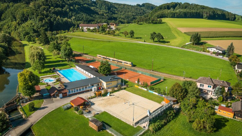 Pielachtalbad Rabenstein, © Markus Haslinger Luftaufnahme eines Freibads mit Pool, Tennisplätzen und Volleyballfeld, umgeben von grünen Feldern und Hügeln.