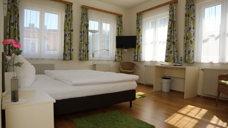 room view, © Gerald Riedler Bright room with bed, desk, TV and several windows with curtains.