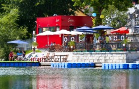 Salettl, © zVg A red building with a terrace by the water, surrounded by parasols and seating.