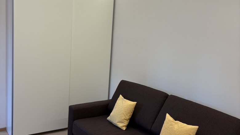 TDR-Haus Morgensonne Wohnzimmer, © Fam Pauker Modernes Wohnzimmer mit braunem Sofa, gelben Kissen und kleinem Tisch mit weißer Tischdecke.