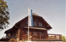 Hainfelder Hütte, © Archiv Stadt Hainfeld Holzhütte mit Fahne auf Terrasse, blauer Himmel.