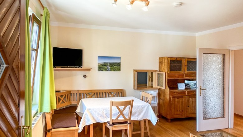 Haus Morgensonne Esszimmer, © Fam. Pauker Esszimmer mit Holzmöbeln, Tisch mit weißer Tischdecke, grünen Vorhängen und Fernseher an der Wand.