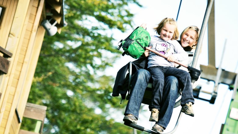 Lift ride up the Muckenkogel, © weinfranz.at Two people on a chair lift, surrounded by trees.