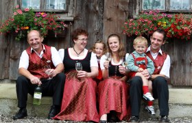 Reikersdorfer's Presshausheuriger, © weinfranz.at Eine Familie in traditioneller Kleidung sitzt vor einem Holzhaus mit Blumen und hält Weingläser.
