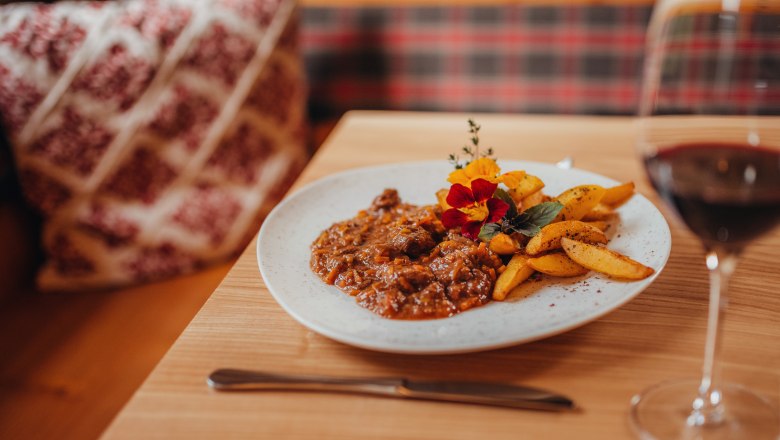 Lamb ragout with thyme potatoes, © Niederösterreich Werbung/Daniela Führer A plate of lamb ragout and thyme potatoes, decorated with edible flowers, on a wooden table. A glass of red wine stands next to it.
