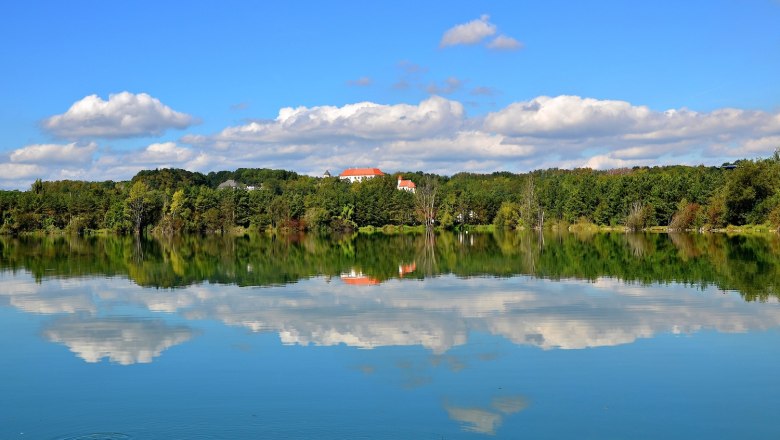 Provincial capital St. Pölten, © SEPA Media, Barbara Seiberl-Stark A lake with a reflection of trees and clouds, in the background a building with a red roof.