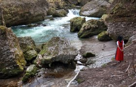 Wonderful - a hike through the gorge, © weinfranz.at A woman in traditional dress stands on the bank of a river in a rocky gorge.