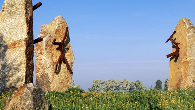Place of the forgotten peoples, © Weinfranz Stone sculptures with metal figures on a meadow under a blue sky.