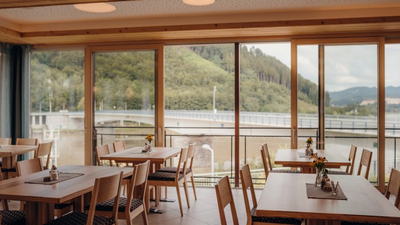 Die Donau und die Stadt Grein immer im Blick, © Niederösterreich Werbung/Daniela Führer Innenansicht eines Gasthofes mit großen Fenstern und Blick auf eine Brücke und bewaldete Hügel.