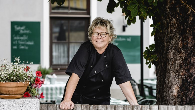 Wirtin Martina Ebner, © Niederösterreich Werbung/David Schreiber Eine Frau in schwarzer Kleidung lehnt an einem Holzzaun, mit Blumen und einem Baum im Hintergrund.