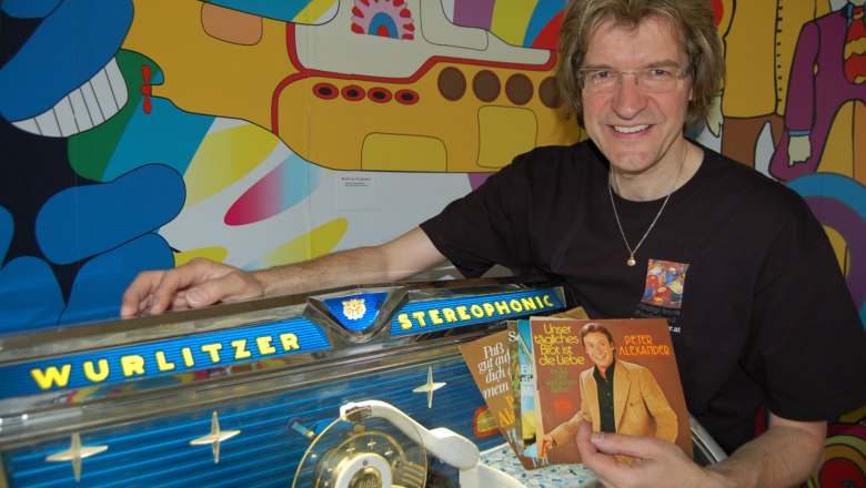 Pindigiland, © Pindigiland Man next to a Wurlitzer jukebox with records in his hand, in front of a colorful background with a yellow submarine.