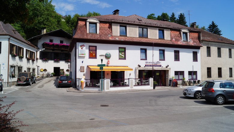 Dirndlhof, © zVg Ein Gebäude mit der Aufschrift 'Dirndlhof' und einer Terrasse mit Sonnenschirm. Autos sind auf dem Parkplatz davor geparkt.
